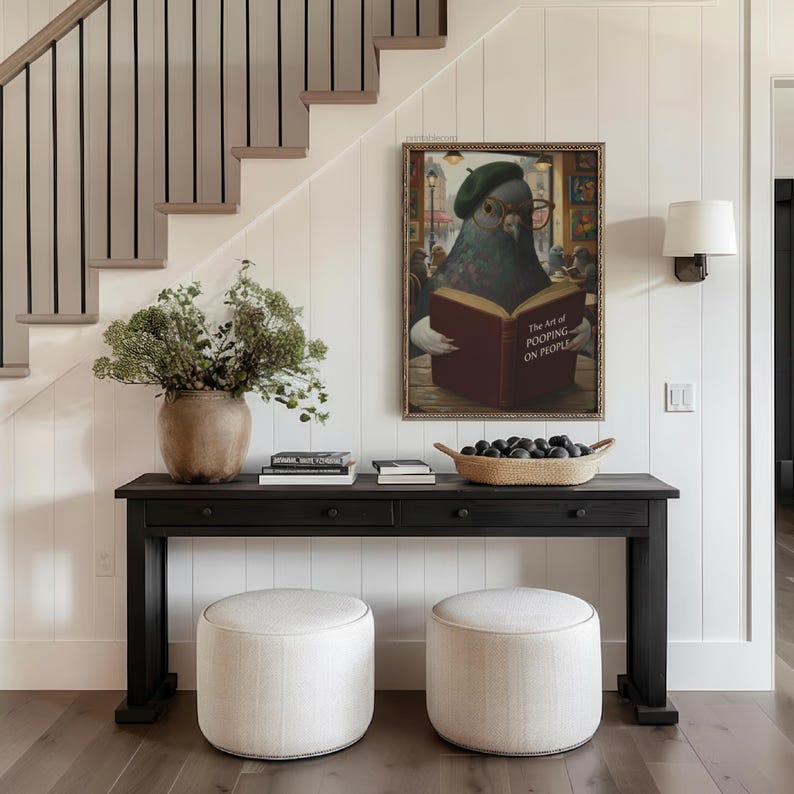 May include: A black console table with two drawers, topped with books, a woven basket, and a large vase with greenery. Two round, upholstered ottomans sit below. A framed artwork depicts a pigeon reading a book with the text "The Art of Pooping on People."