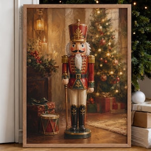 Op de afbeelding: Een ingelijste kunstdruk met een klassieke notenkraker-soldaat in een rood en groen uniform met gouden accenten, staand op een ronde voet. De scène bevat een versierde kerstboom, ingepakte cadeaus en een trommel op de achtergrond.