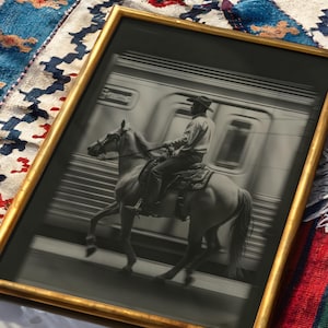 May include: Black and white framed print of a cowboy riding a horse past a train. The print has a gold frame and is set against a colorful patterned textile.