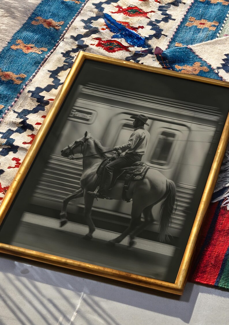 May include: Black and white framed print of a cowboy riding a horse past a train. The print has a gold frame and is set against a colorful patterned textile.