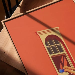 May include: A framed art print featuring a stylized illustration of a window with an arched top. The artwork has a warm, orange background with a window frame in cream and red. A person is reading a book in the window.