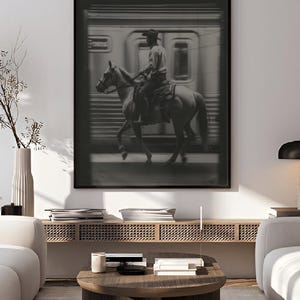 May include: Black and white framed print featuring a cowboy on horseback riding past a train. The image has a vintage aesthetic, with the text "printablecorp" in the top left corner. The artwork is displayed in a modern living room setting.