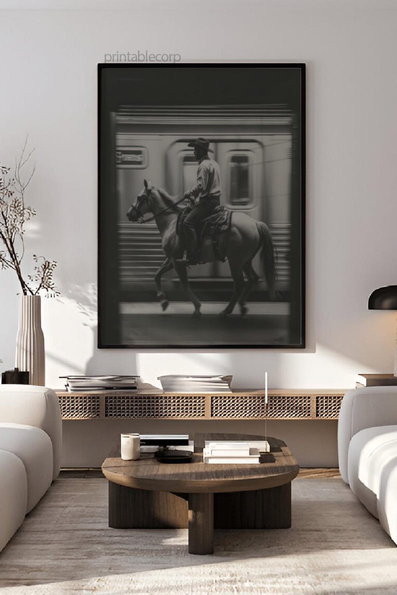 May include: Black and white framed print featuring a cowboy on horseback riding past a train. The image has a vintage aesthetic, with the text "printablecorp" in the top left corner. The artwork is displayed in a modern living room setting.