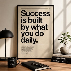 Könnte beinhalten: Ein gerahmter Druck mit dem Text "Success is built by what you do daily" in schwarzer Schrift. Ein schwarzer Becher mit den Worten "Discipline, Focus, Consistency, Success" steht auf einem Holztisch, zusammen mit einem schwarzen Notizbuch, Stift und Büchern.