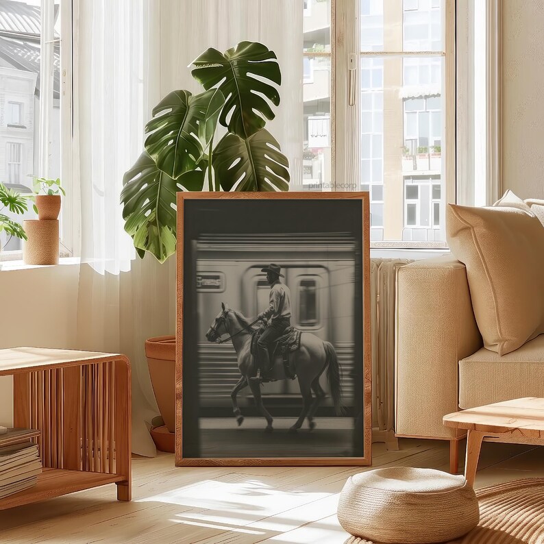 May include: A framed black and white photograph depicts a cowboy riding a horse in front of a train. The image is set in a room with a large window, a potted plant, and wooden furniture. The photograph is in a wooden frame.