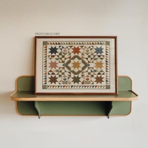 May include: A framed print featuring a geometric quilt pattern with stars and floral motifs in muted earth tones, including brown, green, blue, and orange. The text PRINTABLECORP is visible above the frame, which rests on a green wall shelf.