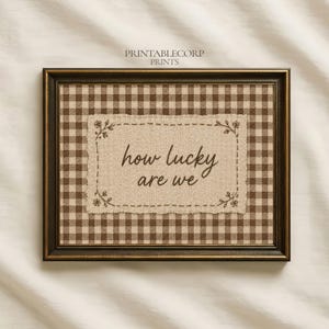 May include: Framed wall art with a brown and tan gingham pattern background. The artwork features a rectangular patch with the phrase "how lucky are we" in a handwritten style. The frame is dark brown.