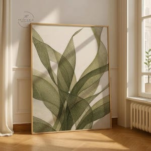 Könnte beinhalten: Ein gerahmter botanischer Druck mit einem abstrakten Design aus sich überlappenden grünen Blättern auf cremefarbenem Hintergrund. Das Kunstwerk wird in einem Raum mit Fenster und Holzboden ausgestellt. Der Druck hat einen hellen Holzrahmen.