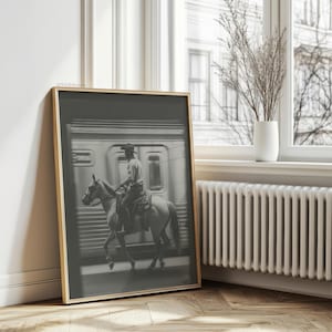 May include: Black and white framed print featuring a cowboy riding a horse in front of a train. The image has a vintage aesthetic, with the cowboy wearing a hat and the horse in motion. The frame is a light wood color.