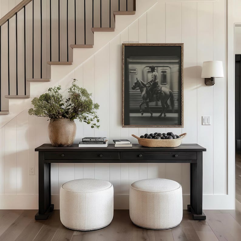 May include: A black console table with two drawers, topped with books, a woven basket of dark spheres, and a large vase of greenery. Two upholstered ottomans sit below. A framed black and white image of a cowboy on a horse hangs above.