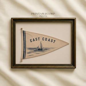 May include: A framed triangular pennant featuring a beige fabric background with two vertical navy blue stripes on the left side. The text "EAST COAST" is printed in navy blue above a navy blue illustration of a sailboat on the water.