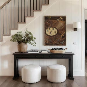 May include: A dark wood console table with two drawers, topped with books, a woven basket of dark spheres, and a large vase of greenery. Two upholstered ottomans sit below. A framed artwork hangs on the white paneled wall.