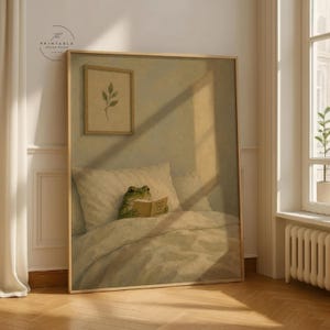 May include: A framed print featuring a frog reading a book in bed. The frog is green and the book is yellow. The bed has white bedding and pillows. A small framed botanical print hangs above the bed. The print is in a gold frame.