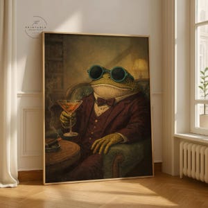 Puede incluir: Impresión enmarcada que presenta una rana elegante con traje burdeos, pajarita y gafas de sol, sosteniendo un cóctel. Un cigarro descansa sobre una pequeña mesa. La obra de arte tiene un estilo vintage y caprichoso, ideal para decoración de pared.