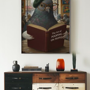 May include: A framed print featuring a pigeon wearing glasses and a beret, reading a book titled "The Art of Pooping on People." The artwork is displayed above a wooden dresser with various decorative items.