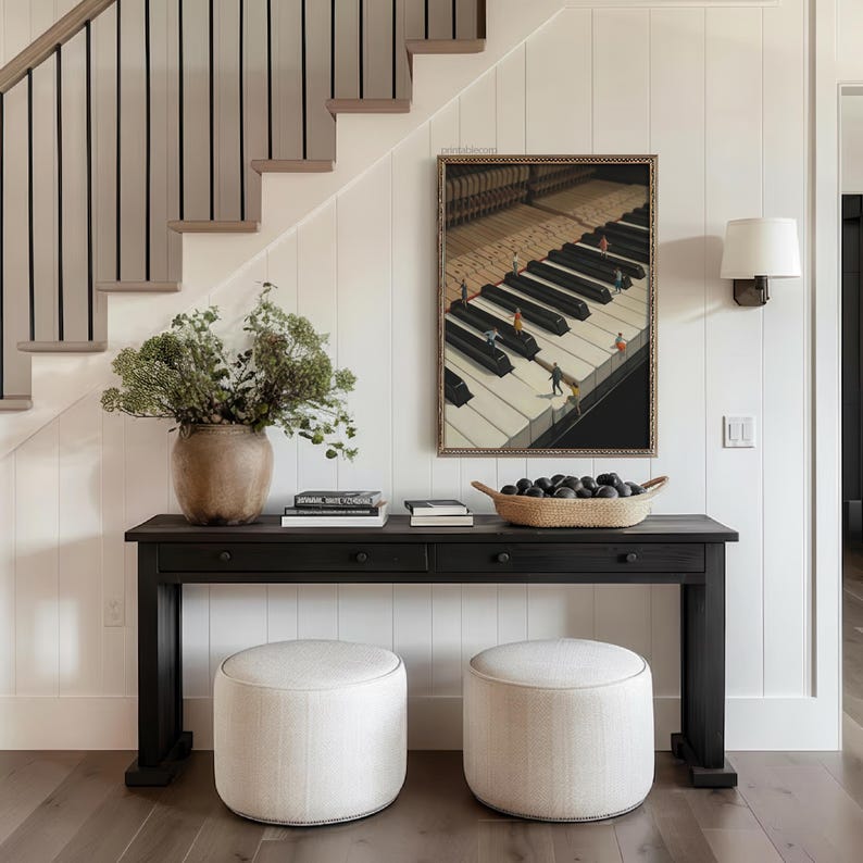 May include: A black console table with two drawers, topped with books, a woven basket of dark objects, and a large vase of greenery. Two round, off-white upholstered ottomans sit in front of the table. A framed artwork of a piano keyboard hangs on the wall.