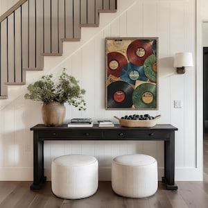 May include: A dark wood console table with two drawers, topped with books, a woven basket, and a large vase of greenery. Two round, upholstered ottomans sit in front of the table. A framed artwork of vintage vinyl records hangs on the wall.