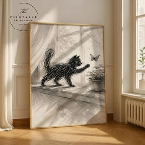 Könnte beinhalten: Schwarz-Weiß-Kunstdruck mit einer Katze und einem Schmetterling. Die Zeichnung zeigt eine schwarze Katze, die nach einem Schmetterling greift, der sich in der Nähe einer Topfpflanze auf einer Fensterbank befindet. Der Druck ist in einem hellen Holzrahmen gerahmt. Logo von The Printable Design Studio.