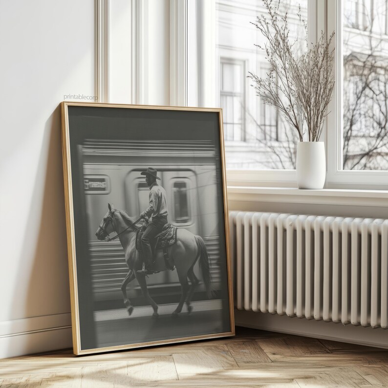 May include: Black and white framed print of a cowboy riding a horse in front of a train. The image is in a gold frame and is leaning against a wall next to a window. The print is a photograph.