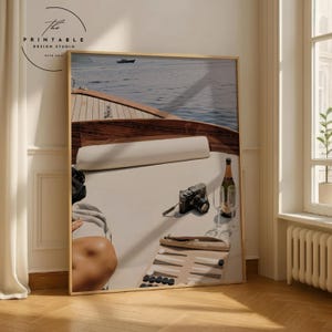 May include: A framed print featuring a boat on the water with a camera, champagne, and backgammon set. The boat has a wooden deck and white cushions. The image has a vintage aesthetic.