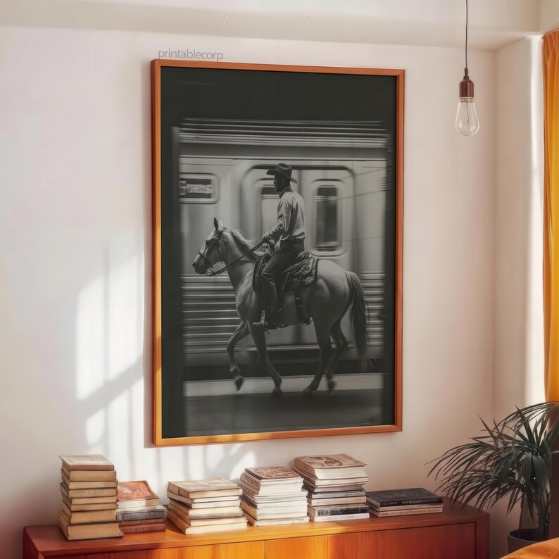 May include: Black and white framed print of a cowboy riding a horse in front of a moving train. The print has a wooden frame and is hanging on a white wall. The print is above a wooden cabinet with books.