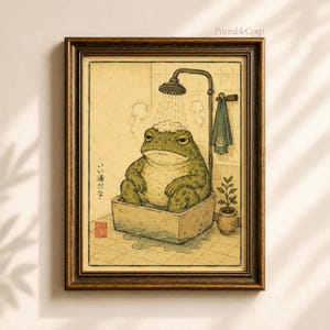 May include: A framed wall art print featuring a green toad sitting in a rectangular stone bathtub under a shower head. The toad has foam on its head. The illustration includes Japanese text, a hanging towel, and a small potted plant in a corner.