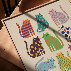 Puede incluir: Una impresi&oacute;n en lienzo enmarcada con una colecci&oacute;n de ilustraciones de gatos coloridos y estilizados. Los gatos se representan en varias poses y patrones, incluyendo rayas, lunares y colores s&oacute;lidos. La impresi&oacute;n tiene un fondo neutro y un marco de madera.