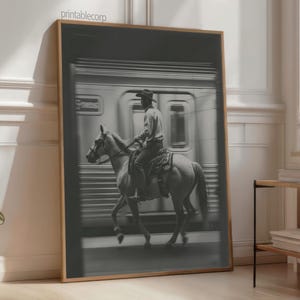 May include: Black and white framed print featuring a cowboy riding a horse in front of a train. The cowboy wears a hat and is seated on a saddle. The image has a vintage aesthetic and is displayed in a room with natural light.