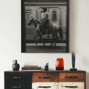 May include: Black and white framed print featuring a cowboy riding a horse in front of a blurred train. The print hangs above a wooden dresser with drawers in black, white, and brown. A red-orange vase and other decorative items are on the dresser.