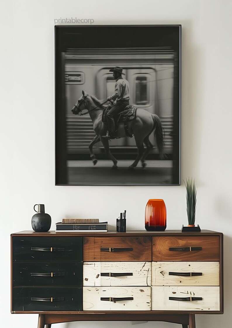 May include: Black and white framed print featuring a cowboy riding a horse in front of a blurred train. The print hangs above a wooden dresser with drawers in black, white, and brown. A red-orange vase and other decorative items are on the dresser.