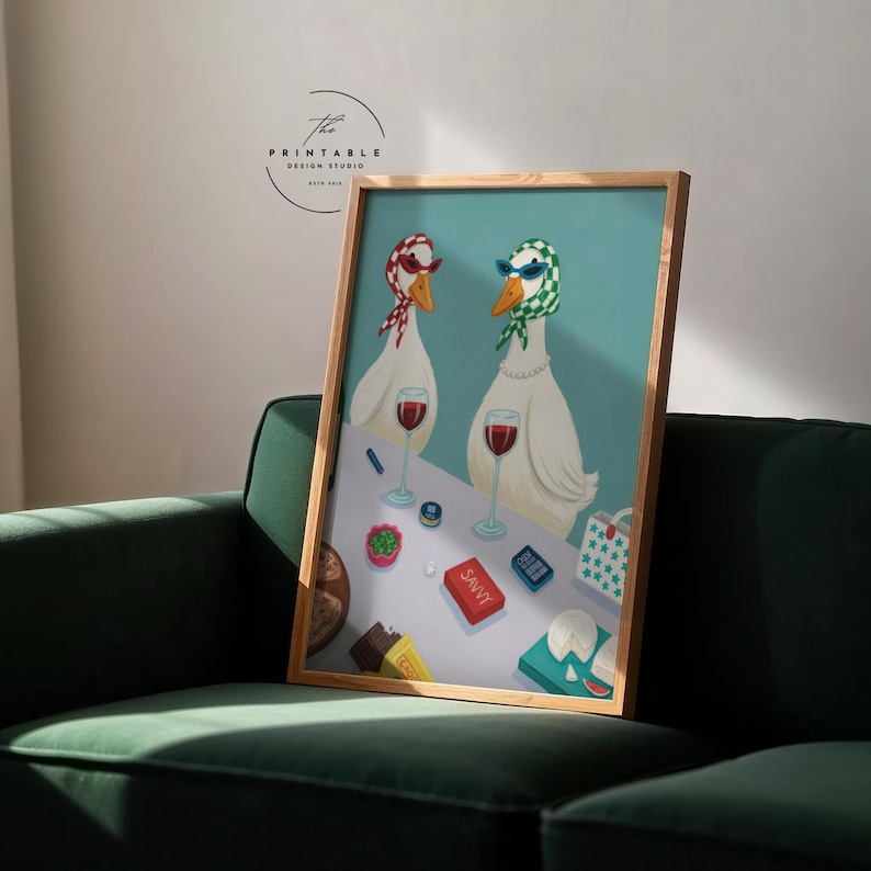 May include: A framed art print featuring two white geese wearing sunglasses and headscarves, standing at a table with wine glasses and snacks. The word "SAVVY" is visible on a red box. The print is in a wooden frame.
