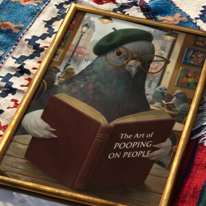 May include: Framed artwork featuring a pigeon wearing a green beret and glasses, reading a book titled "The Art of Pooping on People." The artwork has a gold frame and depicts a Parisian cafe scene.
