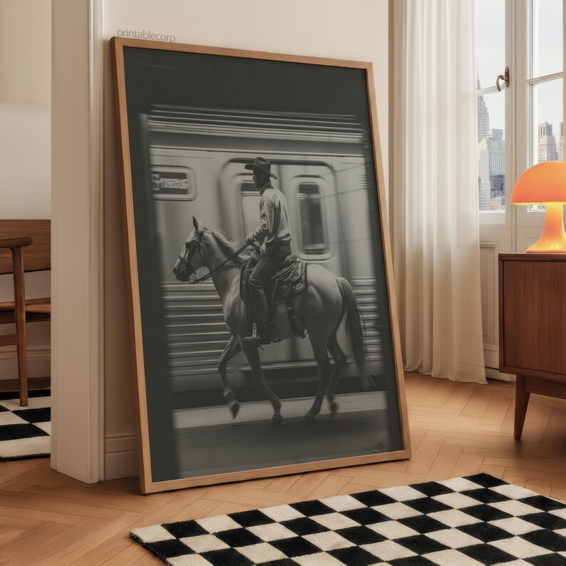 May include: Black and white framed print of a cowboy riding a horse in front of a train. The cowboy is wearing a hat and riding a saddled horse. The train is in the background. The print is leaning against a wall.