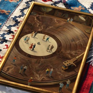 May include: A framed painting depicts a vintage record player with miniature figures dancing on the record's surface. The painting has a gold frame and is set against a patterned rug. The record player's arm and needle are visible.
