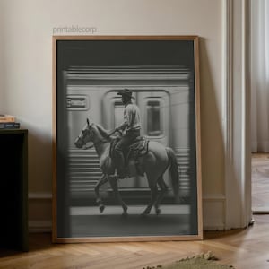 May include: Black and white framed print of a cowboy riding a horse in front of a subway train. The cowboy wears a hat and is seated on a saddle. The image has a vintage, cinematic feel.