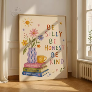 May include: A framed art print with a cream background features colourful illustrations and the words "BE SILLY, BE HONEST, BE KIND." A vase of flowers, a yellow mug, and stacked books are also depicted. The print has a wooden frame.