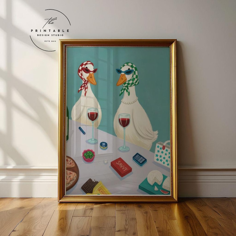 May include: Framed art print featuring two geese wearing sunglasses and scarves, enjoying wine. The table includes pizza, cheese, and a box labeled "SAVVY." The print is in a gold frame, with the text "The Printable Design Studio" in the upper left corner.