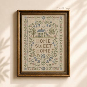 May include: Framed cross-stitch artwork with a house, trees, and floral details in blue, pink, and green. The text "HOME SWEET HOME" is stitched in brown. The frame is dark wood.