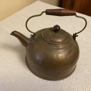 May include: A vintage copper tea kettle with a wooden handle. The kettle has a tarnished finish and a classic design.