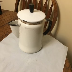 May include: White enamel coffee pot with a black handle and lid. The pot has a spout on the side and a black knob on the lid.