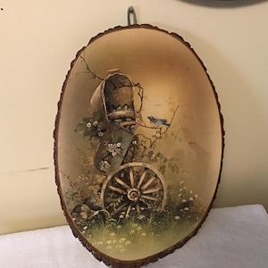 May include: A round wooden slice with a painted scene of a birdhouse with birds, a wheel, and flowers. The background is a light brown with a hint of blue.