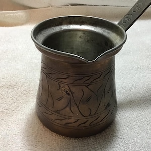 May include: A vintage copper coffee pot with an etched floral design. The pot has a long handle and a spout for easy pouring. The pot is made of copper and has a tarnished finish, giving it a rustic look.