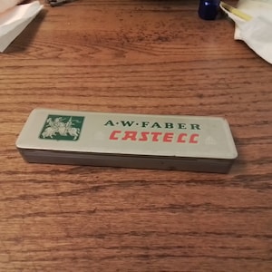 May include: A silver metal box with a green and red label that reads "A.W. Faber Castell". The label features a knight on horseback.