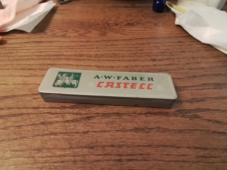 May include: A silver metal box with a green and red label that reads "A.W. Faber Castell". The label features a knight on horseback.