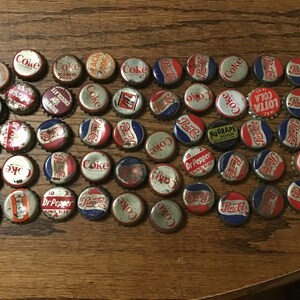 May include: A collection of vintage soda bottle caps, including brands like Coca-Cola, Pepsi-Cola, 7-Up, and Dr. Pepper. The caps are primarily red, white, and blue, with some showing signs of age and wear. The caps are arranged on a wooden surface.