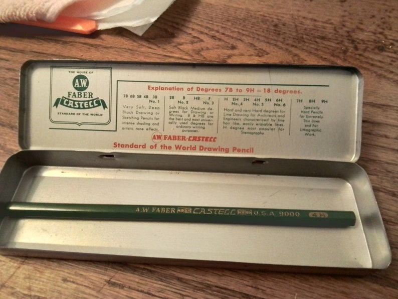 May include: A vintage tin box with a green and white label that reads "A.W. Faber-Castell Standard of the World Drawing Pencil." The label includes a chart explaining the different degrees of hardness for drawing pencils. The box is open and contains a green A.W. Faber-Castell pencil.