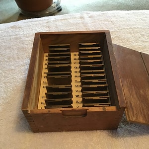 May include: A wooden box with a hinged lid, containing a set of 26 metal letter tiles. The tiles are arranged alphabetically and are labeled with the letters A through Z. The box is made of light brown wood and has a worn finish.