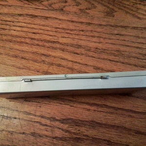 May include: A silver metal rectangular box with a hinged lid and a metal clasp. The box is empty and sitting on a wooden surface.