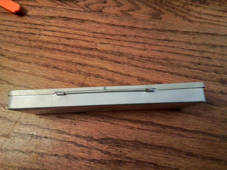 May include: A silver metal rectangular box with a hinged lid and a metal clasp. The box is empty and sitting on a wooden surface.