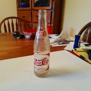 May include: A vintage glass Pepsi-Cola bottle with a red and white label. The bottle is clear and has a textured surface.